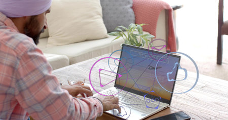 Man wearing purple turban, plaid shirt typing at home desk, laptop showing social icons, copy space....
