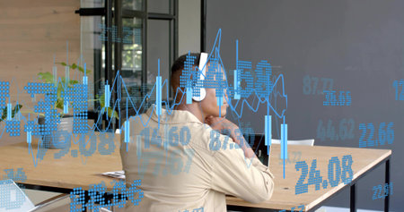 Listening man wearing white-headset and beige shirt in modern office, with laptop and blue charts....