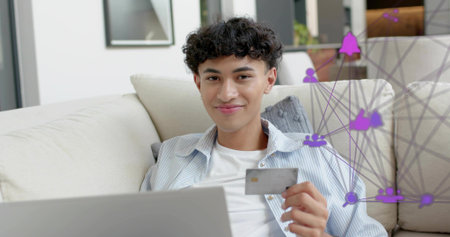 Using laptop and holding metallic card, man wearing blue-striped shirt at home with purple overlay....