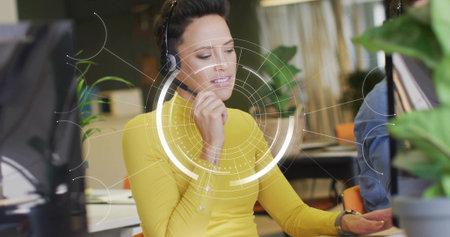 Speaking woman wearing bright yellow top and headset gesturing at screen in open office with HUD....