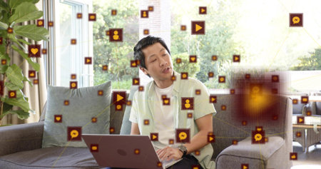 Typing Asian man in green shirt on gray sofa at home, using laptop with floating icons....