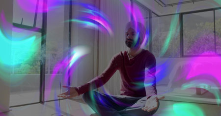 Sitting turbaned Indian man in red shirt meditating at home by tall windows with colorful streaks....