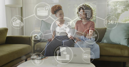 Discussing two women in casual clothes reviewing tech icons at home on sofa, with laptop, tablet....