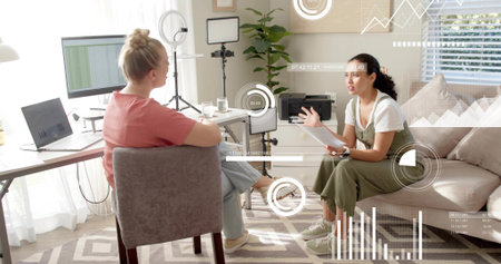 Talking two women wearing coral top, olive overalls in home office, holding papers, laptop....