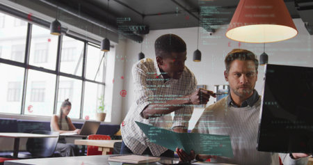 Leaning men pointing at widescreen monitor in open plan office in checked shirt with blue folder....