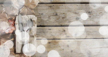 Displaying folded beige napkin holding steel fork and spoon on planks with dried leaves, copy space....