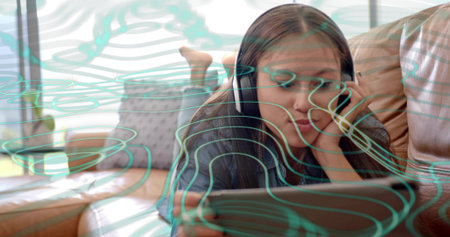 Lying Asian teen wearing dark top, headphones, holding tablet on tan sofa, barefoot near window....