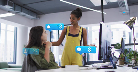 Pointing woman in yellow overalls leaning toward colleague at office, with laptop and notifications....