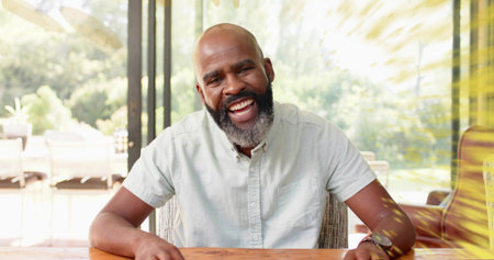Laughing man wearing short-sleeve shirt leaning toward camera at home, wooden table, glass doors....