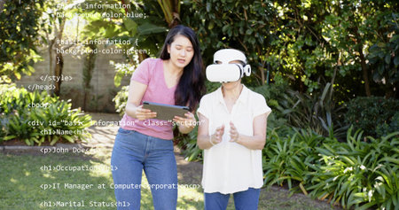 Guiding caregiver holding tablet and leaning toward senior in VR headset in backyard. Outdoor,...