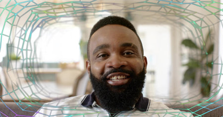 Smiling bearded man wearing zip-up shirt, sitting on sofa in bright living room, polygonal overlay....