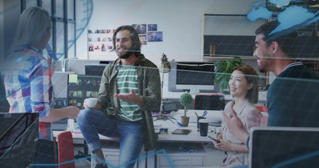 Gesturing bearded man in green shirt leading team discussion at open-plan office, with laptops mugs....
