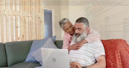 Leaning mature couple in pink blouse and striped shirt viewing laptop on sofa with blue pillow....