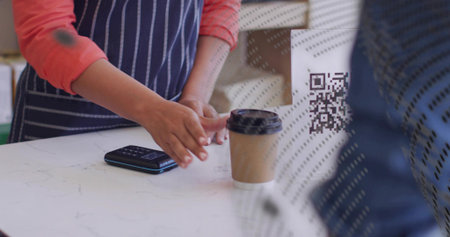 Reaching barista in navy apron and coral top placing cup beside payment device on marble counter....