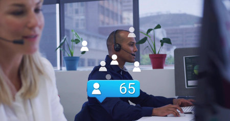 Typing call-center agent wearing navy shirt and headset at open-plan desk, showing blue 65 overlay....
