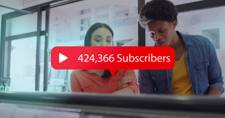 Leaning coworkers checking screen in office, orange blouse, denim shirt showing red 424,366 subs....
