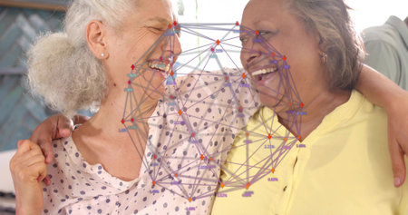 Laughing senior women hugging at home, showing patterned blouse, pale yellow top. Companionship,...