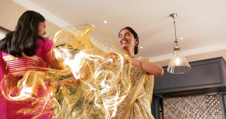 Dancing woman in pale yellow gold saree twirling at home kitchen, with pink saree, pendant lamp....