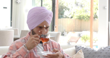 Sipping turbaned man in pink plaid shirt, holding glass teacup, saucer at home, sliding doors open....