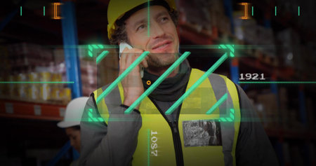 Speaking supervisor in yellow hard hat and vest using smartphone in warehouse with AR HUD overlays....