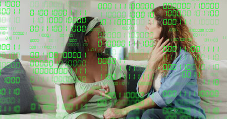 Holding hands, two women sharing talk on sofa at home, wearing headband, with green binary overlay....