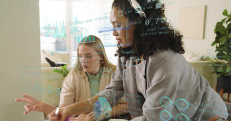 Analyzing two women pointing at holographic charts in home office lounge, wearing blazers....
