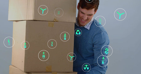 Holding three taped cardboard boxes, man in blue sweater bracing in studio with green digital icons....