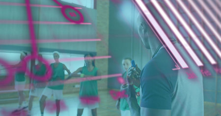 Leading coach in polo holding timer, briefing green-vested team in gym under pink pane, copy space....