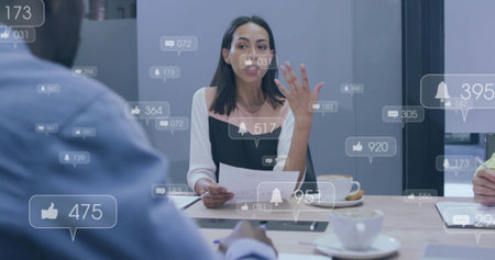 Speaking woman in black top with white sleeves, holding documents at meeting table, social overlays....