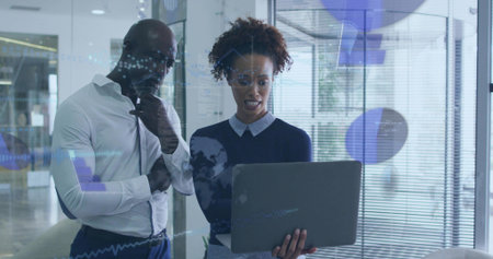 Reviewing man in white shirt, woman in dark sweater holding laptop at modern office glass partition....