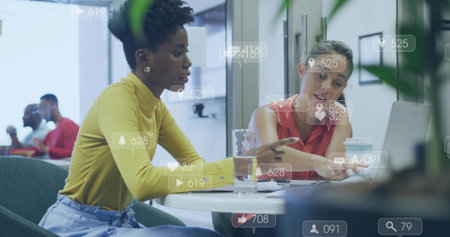 Leaning women wearing mustard and coral tops pointing at laptop with social overlays in office....