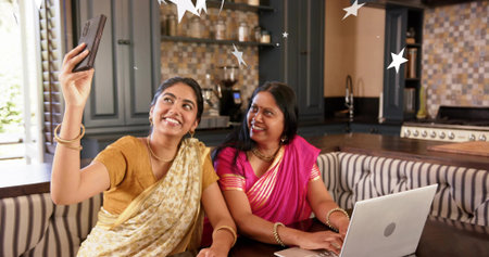 Smiling daughter holding smartphone, mother watching laptop at kitchen nook in sarees. Family, home,...