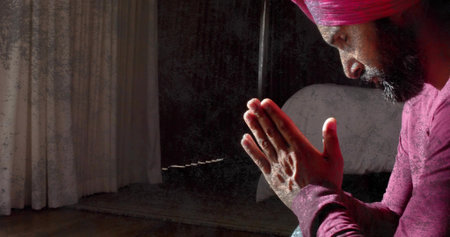 Praying mid-adult man wearing pink turban and maroon shirt, sitting right in dim room, copy space. Contemplation, meditation, solitude, serenity, stillness, sidelit, woodenの写真素材