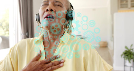 Listening senior woman holding hand on chest at home, wearing headset, pale blouse. Portrait,...