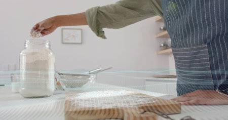 Scooping flour from glass jar, woman in pinstripe apron, green shirt at kitchen counter, copy space....