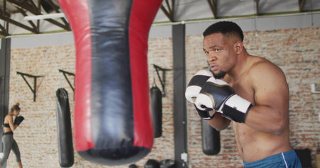 Training shirtless boxer in blue shorts and boxing gloves guarding gym heavy bag. Athlete, fitness,...