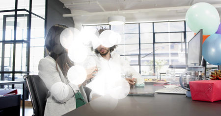 Chatting white-shirted man and blazer-clad woman holding mug in office, with laptop and balloons....