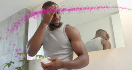 Man in gray tank top applying cream to forehead at bathroom mirror, holding small white jar....