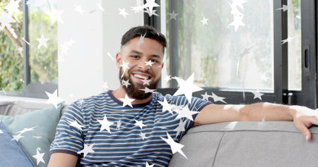 Sitting man wearing striped-tee smiling on sofa at home, with black-windows, star-stickers. Adult,...