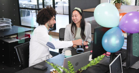 Reaching coworkers wearing white shirt, light blazer working at office desk with laptop, balloons....