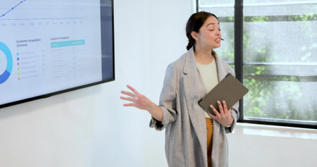 Presenting presenter in gray blazer speaking, gesturing in meeting room with tablet, showing charts....
