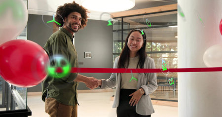 Shaking hands green-shirted man and woman in gray blazer in office lobby, red ribbon and balloons....