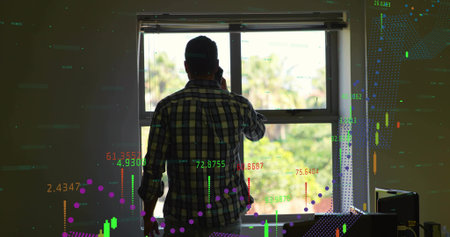 Holding smartphone, mid adult man in plaid shirt and cap watching market charts at home window....