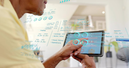Holding tablet, senior woman wearing pale yellow top tapping touchscreen at home, with AR graphics....