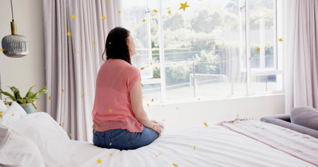 Sitting Asian woman wearing pink top and jeans gazing out window, with bed, pastel throw....