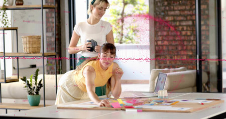 Working two women wearing yellow top, arranging sticky notes at large work table in studio, laptop....