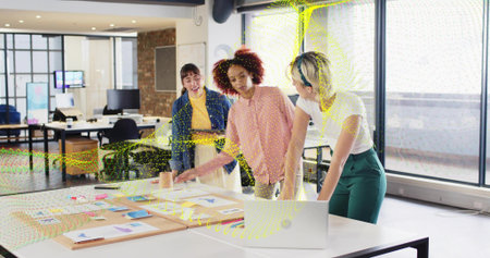 Reviewing mockups and layouts, three women leaning over white table in modern office with laptop....