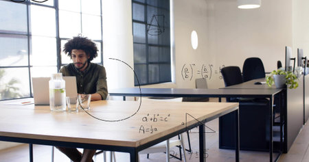 Working man in olive-green shirt typing on laptop at open office desk, with pitcher and equations....