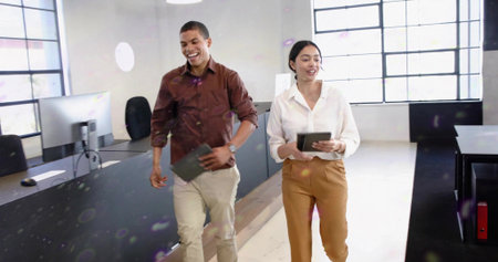 Walking man in burgundy shirt and woman in white blouse holding tablets, speaking in open office....