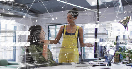 Gesturing standing woman yellow overalls speaking to colleague in modern office with laptop monitor. Collaboration, teamwork, openplan, workspace, diagrams, daylight, sleekの写真素材